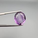 A 11.05-carat natural purple Amethyst (Katela) from Brazil, featuring a brilliant oval faceted cut with exceptional clarity and deep violet saturation, shown in jeweler's tweezers.