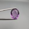 ​A 11.91-carat natural Brazilian Amethyst (Katela) gemstone, featuring a brilliant oval faceted cut with deep purple saturation and excellent clarity, held in jeweler's tweezers.