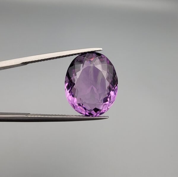 ​A 11.91-carat natural Brazilian Amethyst (Katela) gemstone, featuring a brilliant oval faceted cut with deep purple saturation and excellent clarity, held in jeweler's tweezers.