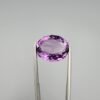 ​A 11.91-carat natural Brazilian Amethyst (Katela) gemstone, featuring a brilliant oval faceted cut with deep purple saturation and excellent clarity, held in jeweler's tweezers.