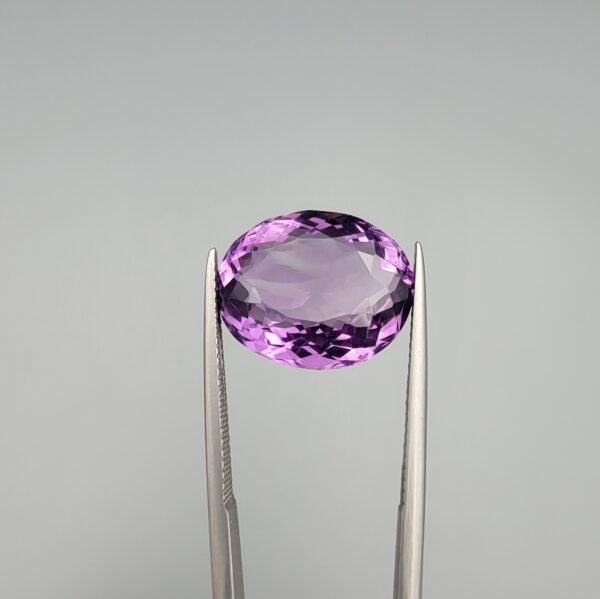 ​A 11.91-carat natural Brazilian Amethyst (Katela) gemstone, featuring a brilliant oval faceted cut with deep purple saturation and excellent clarity, held in jeweler's tweezers.