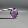 ​A 11.91-carat natural Brazilian Amethyst (Katela) gemstone, featuring a brilliant oval faceted cut with deep purple saturation and excellent clarity, held in jeweler's tweezers.