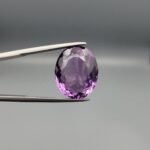 ​A 11.91-carat natural Brazilian Amethyst (Katela) gemstone, featuring a brilliant oval faceted cut with deep purple saturation and excellent clarity, held in jeweler's tweezers.