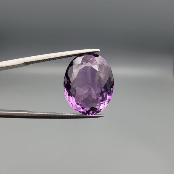 ​A 11.91-carat natural Brazilian Amethyst (Katela) gemstone, featuring a brilliant oval faceted cut with deep purple saturation and excellent clarity, held in jeweler's tweezers.
