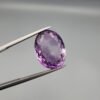 A large 13.51-carat natural Brazilian Amethyst (Katela) gemstone, showcasing a brilliant oval faceted cut with intense royal purple color and high transparency, held in jeweler's tweezers.