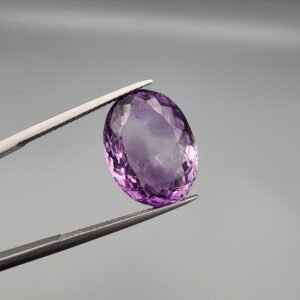 A large 13.51-carat natural Brazilian Amethyst (Katela) gemstone, showcasing a brilliant oval faceted cut with intense royal purple color and high transparency, held in jeweler's tweezers.