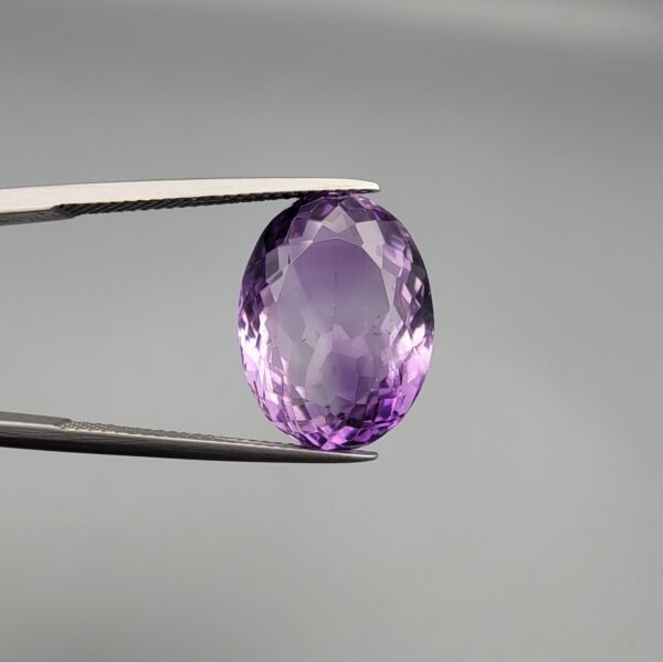 ​A 14-carat natural Brazilian Amethyst (Katela) gemstone, featuring a brilliant oval faceted cut with deep violet saturation and exceptional eye-clean clarity, held in jeweler's tweezers.