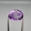 ​A 14-carat natural Brazilian Amethyst (Katela) gemstone, featuring a brilliant oval faceted cut with deep violet saturation and exceptional eye-clean clarity, held in jeweler's tweezers.