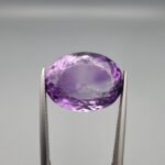 ​A 14-carat natural Brazilian Amethyst (Katela) gemstone, featuring a brilliant oval faceted cut with deep violet saturation and exceptional eye-clean clarity, held in jeweler's tweezers.