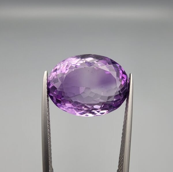​A 14-carat natural Brazilian Amethyst (Katela) gemstone, featuring a brilliant oval faceted cut with deep violet saturation and exceptional eye-clean clarity, held in jeweler's tweezers.