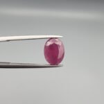 ​A 4.61-carat natural Mozambique Ruby (Yaqoot) gemstone, featuring a brilliant oval faceted cut with a rich pigeon-blood red hue and excellent transparency, held in jeweler's tweezers.