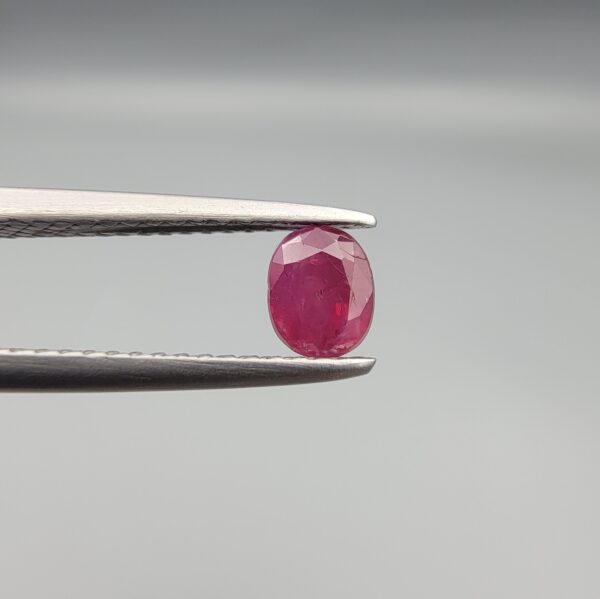 ​A 0.72-carat natural red Ruby (Yaqoot) from Kabul, Afghanistan, featuring an oval cut with a distinct yellow mountain inclusion on the back and a fine natural crack line on the front, held in jeweler's tweezers.