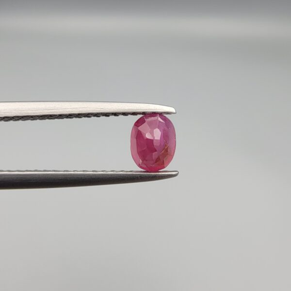 ​A 0.72-carat natural red Ruby (Yaqoot) from Kabul, Afghanistan, featuring an oval cut with a distinct yellow mountain inclusion on the back and a fine natural crack line on the front, held in jeweler's tweezers.
