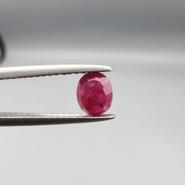 ​A 0.51-carat natural red Ruby (Yaqoot) from Kabul, Afghanistan, featuring an oval faceted cut with a distinct black mountain inclusion on the back side, held in jeweler's tweezers.