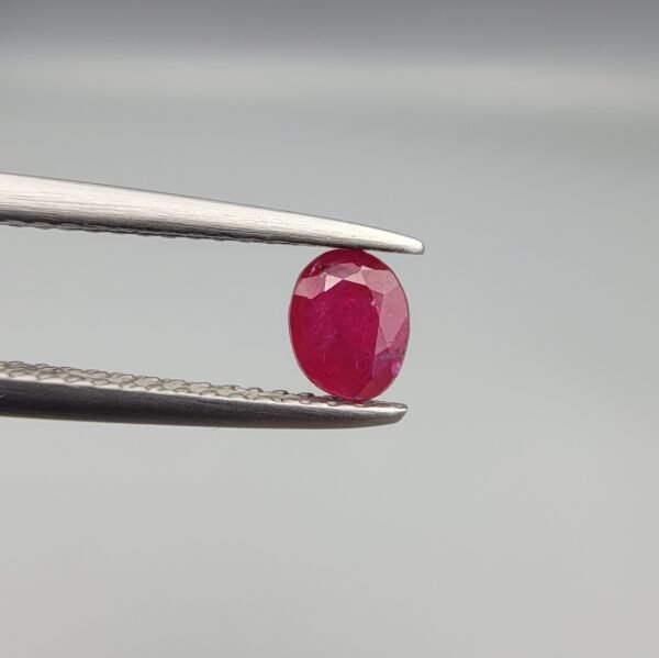 ​A 0.45-carat natural red Ruby (Yaqoot) from Kabul, Afghanistan, featuring an oval faceted cut with visible black mountain mineral inclusions on both the front and reverse sides, held in jeweler's tweezers.