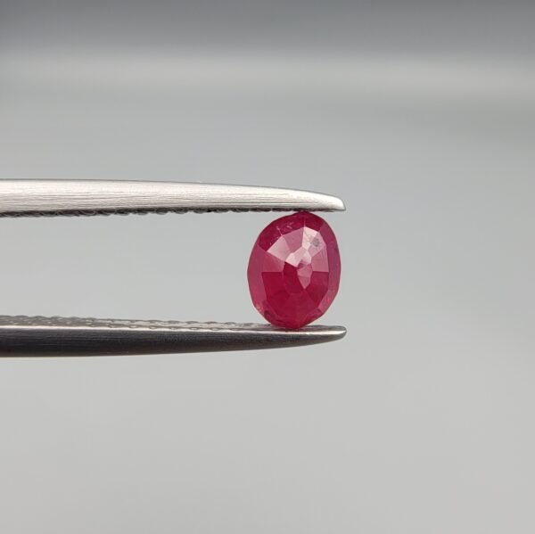 ​A 0.45-carat natural red Ruby (Yaqoot) from Kabul, Afghanistan, featuring an oval faceted cut with visible black mountain mineral inclusions on both the front and reverse sides, held in jeweler's tweezers.