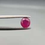 ​A 0.62-carat natural red Ruby (Yaqoot) from Kabul, Afghanistan, featuring a round cut with a natural black mountain inclusion on the back and tiny organic holes on the front, held in jeweler's tweezers.