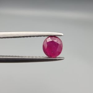 ​A 0.62-carat natural red Ruby (Yaqoot) from Kabul, Afghanistan, featuring a round cut with a natural black mountain inclusion on the back and tiny organic holes on the front, held in jeweler's tweezers.