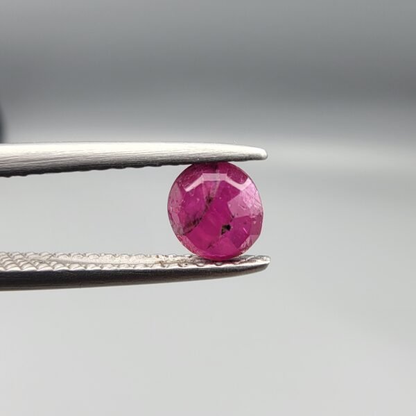 ​A 0.62-carat natural red Ruby (Yaqoot) from Kabul, Afghanistan, featuring a round cut with a natural black mountain inclusion on the back and tiny organic holes on the front, held in jeweler's tweezers.
