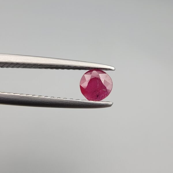 A 0.39-carat natural red Ruby (Yaqoot) from Kabul, Afghanistan, featuring a round brilliant cut with a natural mountain hole on the back and a tiny black inclusion on the front, held in jeweler's tweezers.