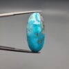 A large 30.79-carat natural blue-green Turquoise (Feroza) from Kerman, Iran, featuring an oval cabochon cut with golden pyrite matrix and natural organic holes on both sides, held in jeweler's tweezers.