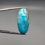 A large 30.79-carat natural blue-green Turquoise (Feroza) from Kerman, Iran, featuring an oval cabochon cut with golden pyrite matrix and natural organic holes on both sides, held in jeweler's tweezers.