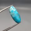 A large 30.79-carat natural blue-green Turquoise (Feroza) from Kerman, Iran, featuring an oval cabochon cut with golden pyrite matrix and natural organic holes on both sides, held in jeweler's tweezers.