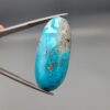 A large 30.79-carat natural blue-green Turquoise (Feroza) from Kerman, Iran, featuring an oval cabochon cut with golden pyrite matrix and natural organic holes on both sides, held in jeweler's tweezers.