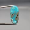 A large 30.79-carat natural blue-green Turquoise (Feroza) from Kerman, Iran, featuring an oval cabochon cut with golden pyrite matrix and natural organic holes on both sides, held in jeweler's tweezers.