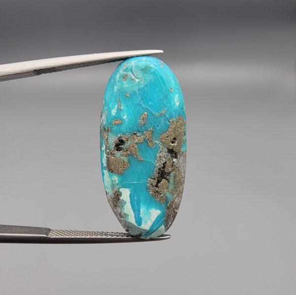 A large 30.79-carat natural blue-green Turquoise (Feroza) from Kerman, Iran, featuring an oval cabochon cut with golden pyrite matrix and natural organic holes on both sides, held in jeweler's tweezers.