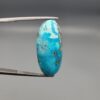 A large 30.79-carat natural blue-green Turquoise (Feroza) from Kerman, Iran, featuring an oval cabochon cut with golden pyrite matrix and natural organic holes on both sides, held in jeweler's tweezers.