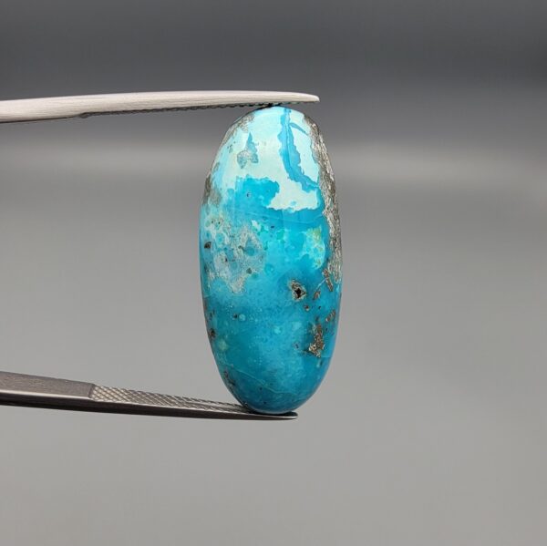 A large 30.79-carat natural blue-green Turquoise (Feroza) from Kerman, Iran, featuring an oval cabochon cut with golden pyrite matrix and natural organic holes on both sides, held in jeweler's tweezers.