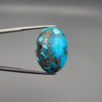 A massive 34.80-carat natural blue-green Turquoise (Feroza) from Kerman, Iran, featuring an oval cabochon cut with a distinct golden pyrite matrix, held in jeweler's tweezers.