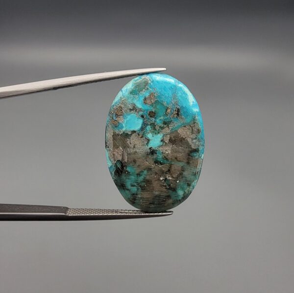 A massive 34.80-carat natural blue-green Turquoise (Feroza) from Kerman, Iran, featuring an oval cabochon cut with a distinct golden pyrite matrix, held in jeweler's tweezers.