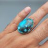A massive 34.80-carat natural blue-green Turquoise (Feroza) from Kerman, Iran, featuring an oval cabochon cut with a distinct golden pyrite matrix, held in jeweler's tweezers.