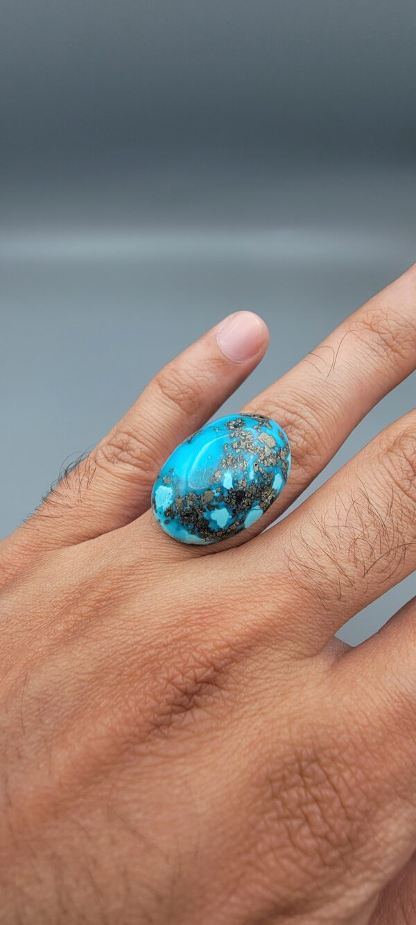 A massive 34.80-carat natural blue-green Turquoise (Feroza) from Kerman, Iran, featuring an oval cabochon cut with a distinct golden pyrite matrix, held in jeweler's tweezers.