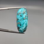 ​A large 36.53-carat natural blue-green Turquoise (Feroza) from Kerman, Iran, featuring a high-dome oval cabochon cut with a rich golden pyrite matrix, held in jeweler's tweezers.
