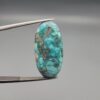 ​A large 36.53-carat natural blue-green Turquoise (Feroza) from Kerman, Iran, featuring a high-dome oval cabochon cut with a rich golden pyrite matrix, held in jeweler's tweezers.