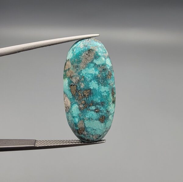 ​A large 36.53-carat natural blue-green Turquoise (Feroza) from Kerman, Iran, featuring a high-dome oval cabochon cut with a rich golden pyrite matrix, held in jeweler's tweezers.