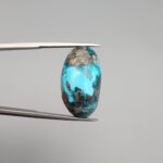 A 25.38-carat natural blue-green Turquoise (Feroza) from Kerman, Iran, featuring an oval cabochon cut with a shimmering golden pyrite matrix, held in jeweler's tweezers.