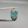 A 25.38-carat natural blue-green Turquoise (Feroza) from Kerman, Iran, featuring an oval cabochon cut with a shimmering golden pyrite matrix, held in jeweler's tweezers.