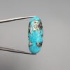​A 28.53-carat natural blue-green Turquoise (Feroza) from Kerman, Iran, featuring an oval cabochon cut with a golden pyrite matrix and natural organic holes on the front and back, held in jeweler's tweezers.