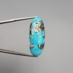 ​A 28.53-carat natural blue-green Turquoise (Feroza) from Kerman, Iran, featuring an oval cabochon cut with a golden pyrite matrix and natural organic holes on the front and back, held in jeweler's tweezers.