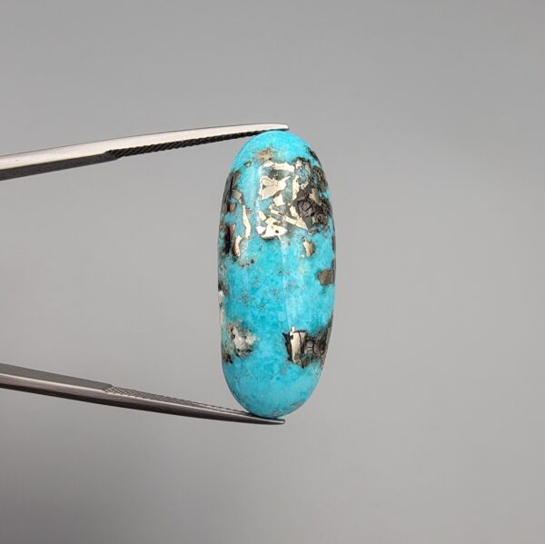 ​A 28.53-carat natural blue-green Turquoise (Feroza) from Kerman, Iran, featuring an oval cabochon cut with a golden pyrite matrix and natural organic holes on the front and back, held in jeweler's tweezers.