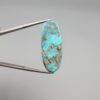 ​A 28.53-carat natural blue-green Turquoise (Feroza) from Kerman, Iran, featuring an oval cabochon cut with a golden pyrite matrix and natural organic holes on the front and back, held in jeweler's tweezers.