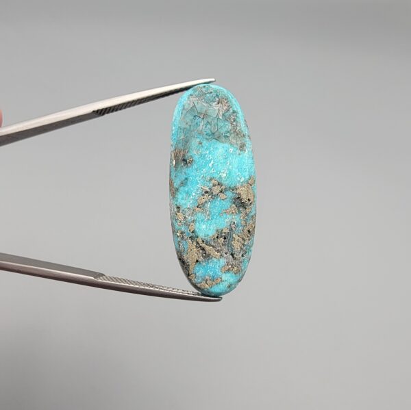 ​A 28.53-carat natural blue-green Turquoise (Feroza) from Kerman, Iran, featuring an oval cabochon cut with a golden pyrite matrix and natural organic holes on the front and back, held in jeweler's tweezers.