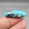 ​A 28.53-carat natural blue-green Turquoise (Feroza) from Kerman, Iran, featuring an oval cabochon cut with a golden pyrite matrix and natural organic holes on the front and back, held in jeweler's tweezers.