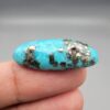 ​A 28.53-carat natural blue-green Turquoise (Feroza) from Kerman, Iran, featuring an oval cabochon cut with a golden pyrite matrix and natural organic holes on the front and back, held in jeweler's tweezers.