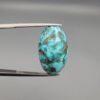 A 16.41-carat natural blue-green Turquoise (Feroza) from Kerman, Iran, featuring an oval cabochon cut with white quartz matrix and natural organic holes on both sides, held in jeweler's tweezers.