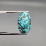 A 16.41-carat natural blue-green Turquoise (Feroza) from Kerman, Iran, featuring an oval cabochon cut with white quartz matrix and natural organic holes on both sides, held in jeweler's tweezers.